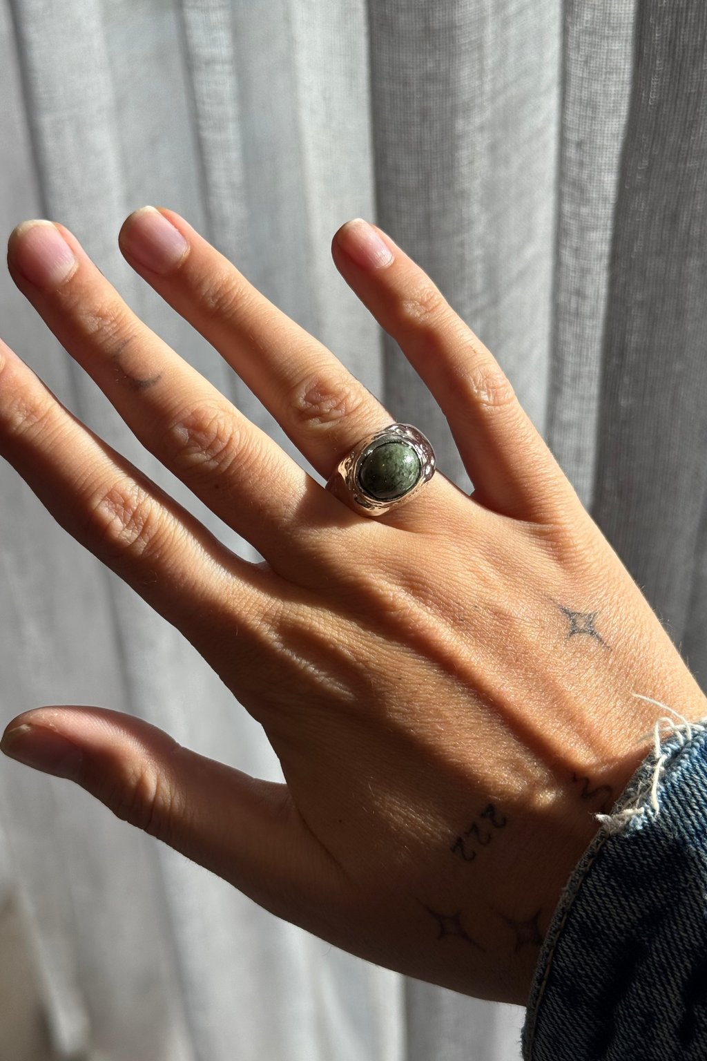 A close-up of a hand wearing a silver ring with a round green stone, set against a soft gray curtain background.